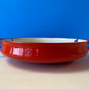 May include: A red enamel cast iron baking dish with two handles. The dish has a white interior and is made of durable cast iron.