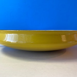 Puede incluir: Una sartén de hierro fundido esmaltado amarillo con dos asas. La sartén es redonda y tiene un fondo ligeramente curvado.