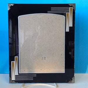 May include: A black picture frame with a gold and white geometric design. The frame has a beveled edge and a clear glass front.