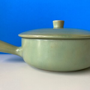 May include: A light green ceramic soup bowl with a lid and a handle. The bowl is round with a curved handle on one side. The lid has a small knob. The bowl is on a white surface with a blue background.