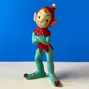 Puede incluir: Una figurita de elfo de cerámica vintage con un gorro rojo y pantalones azules. El elfo tiene una expresión caprichosa y está de pie con los brazos cruzados.
