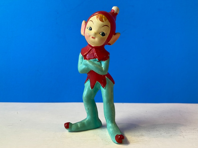 Op de afbeelding: Een vintage keramiek elfenfiguur met een rode muts en blauwe broek. De elf heeft een speelse uitdrukking en staat met gekruiste armen.