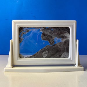 May include: A white, rectangular sand art frame with a stand. The frame contains a landscape scene with black and gray sand against a blue background. The frame is made of white plastic.