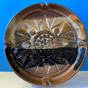 May include: A brown ceramic ashtray with a black and white geometric design. The ashtray has a circular shape and a raised rim.