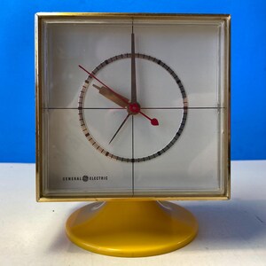 Può includere: Un orologio vintage General Electric con quadrante quadrato e cornice color oro. L'orologio ha uno sfondo bianco con lancette nere e rosse e una base gialla. Il quadrante ha un design circolare con un motivo a reticolo.