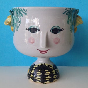 May include: A white ceramic planter shaped like a face with green and yellow painted details. The planter has a wide bowl and a round base.