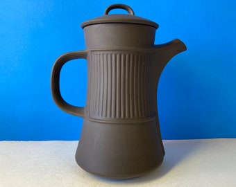 Op de afbeelding: Een donkerbruine keramische koffiepot met een matte afwerking, voorzien van een verticaal geribbelde textuur op het centrale gedeelte, een gebogen handvat, een brede tuit en een bijpassend deksel met een kleine boogvormige handgreep bovenop.