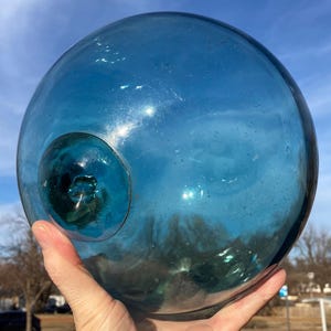 Pode incluir: Um grande flutuador de vidro azul redondo com uma abertura circular menor. O flutuador é segurado em uma mão contra um fundo de céu azul e árvores. O vidro tem uma leve textura e reflete a luz.