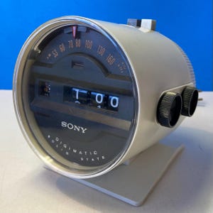 May include: A vintage Sony Digimatic solid state alarm clock. The clock has a cream-colored body with a clear plastic face. The clock face displays the time and radio frequency. Two black knobs are on the side.