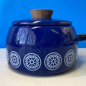 Puede incluir: Una olla de esmalte azul profundo con una tapa a juego que presenta un pomo de madera de color claro. El exterior de la olla está decorado con un patrón repetitivo de motivos circulares blancos, cada uno con un diseño simétrico y detallado.