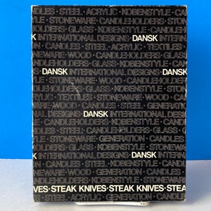 May include: A black and white box with the word "DANSK" repeated multiple times. The box lists various product categories such as "STEAK KNIVES", "CANDLES", and "STONEWARE". The box is on a white surface against a blue background.