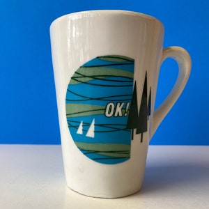 May include: White ceramic mug with a curved handle. The mug features a design with a blue and green circular graphic, with the word "OK!" and stylized trees. The mug is a standard size for coffee or tea.