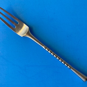 Puede incluir: Un tenedor plateado con tres púas, sobre un fondo azul. El mango tiene un diseño texturizado y estriado. Este utensilio es probablemente para servir o comer.