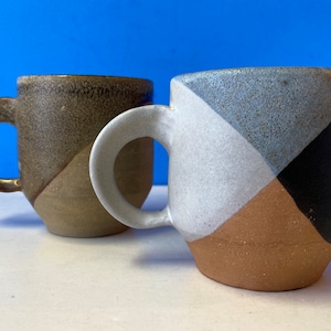 Puede incluir: Dos tazas de cerámica con diseños geométricos. Una taza es marrón, blanca y negra. La otra taza es azul, blanca y marrón.