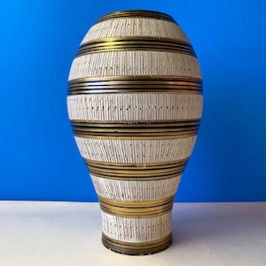 Puede incluir: Un jarrón de cerámica blanco con rayas negras y doradas. El jarrón tiene un cuerpo redondeado y un cuello estrecho.