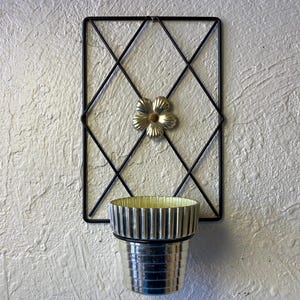 May include: A black metal wall planter with a diamond pattern and a gold flower design. A silver planter sits inside the wall planter.