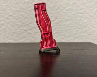 May include: A small, metallic red object with a black base. The object has a rectangular shape with a series of parallel lines and a curved design. The object is angled slightly, resting on a dark surface.