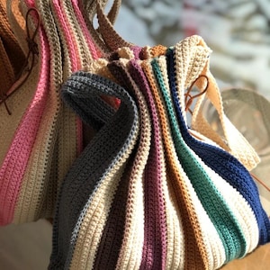 May include: A selection of handmade crochet tote bags with vertical stripes in shades of pink, beige, blue, and brown. Each bag has a loop handle and a leather strap detail. The bags are made from soft, textured yarn, showcasing a variety of colours.