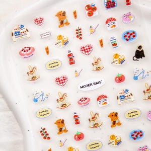 Op de afbeelding: Een vel transparante stickers met diverse kleurrijke illustraties. Ontwerpen omvatten een schommelpaard, een beer, een taart, een tomaat, een hart en een zwarte kat. De tekst "MOOER BMS" is ook aanwezig.