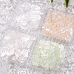 May include: Four clear plastic containers filled with decorative flakes in white, peach, and pale green hues. The containers are arranged on a textured, white surface, creating a visually appealing display for crafts or embellishments.