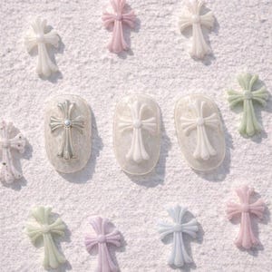 May include: An assortment of decorative cross nail art in various pastel colors, including pink, green, and blue. Some crosses are applied to manicured nails, while others are scattered on a white surface. The crosses have a detailed, ornate design.