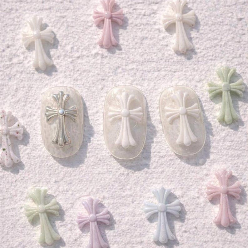 May include: An assortment of decorative cross nail art in various pastel colors, including pink, green, and blue. Some crosses are applied to manicured nails, while others are scattered on a white surface. The crosses have a detailed, ornate design.