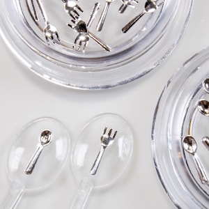 Op de afbeelding: Luchtfoto van miniatuur zilveren vorken en lepels gerangschikt op heldere, ronde, plastic serveerschalen. De bestekken zijn verspreid over de schalen en op het witte oppervlak, waardoor een speelse, grillige esthetiek ontstaat. Ideaal voor poppenhuizen.