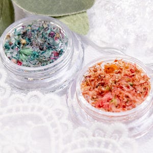 May include: Two small, clear jars filled with colourful, dried floral elements. One jar contains a mix of green, blue, and purple pieces, while the other holds orange and pink fragments. The jars sit on a white, lace-textured surface.