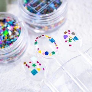 May include: Close-up of nail art supplies. Two clear jars filled with colourful glitter and three clear plastic applicators decorated with various shapes of glitter. The glitter includes blue, green, yellow, and purple colours.