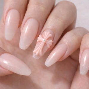 May include: A hand with long, light pink, coffin-shaped acrylic nails. One nail features a 3D pink cross with a pearl center. The nails have a glittery white tip.