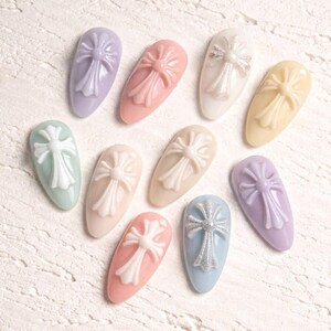 May include: A collection of ten oval-shaped nail art pieces in various pastel colors, each featuring a raised, ornate cross design. Some crosses have a glittery finish, adding a touch of sparkle to the nail art.