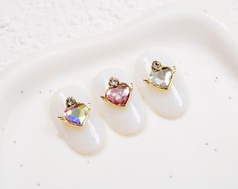 以下が含まれることがあります： イリデセント、ピンク、クリアカラーのハート型宝石をあしらった3つのネイルアートデザイン。それぞれにゴールドカラーのフレームと上部に小さな宝石が付いています。白い楕円形のネイルの形にデザインされ、白い表面に配置されています。