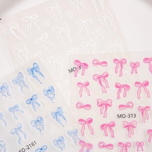 May include: Sheets of nail art stickers featuring bow designs in pink, blue, and white. The pink bows have the text "MO-313" printed on the sheet. The blue bows have the text "SD-2161" printed on the sheet. The bows are in various sizes.