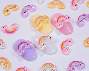 May include: A selection of rainbow nail art decorations. The rainbow designs have smiling cloud accents and are set against a white background. The nail art decorations are in various colours, including yellow, pink, and purple.