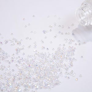 May include: A clear glass bottle, lying on its side, with many small, iridescent crystals scattered across a white surface. The crystals glisten, creating a sparkling effect. The bottle has a textured design.