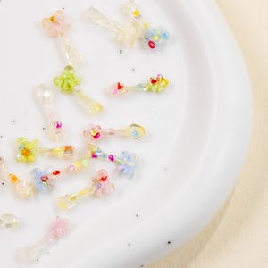 May include: A collection of small, colorful, flower-shaped embellishments with clear stems, scattered on a white plate. The flowers feature various pastel colors, including pink, yellow, blue, and green, with small, sparkling accents.