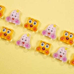 May include: A collection of colorful, cartoon-style charms. The charms feature two distinct designs: one is a yellow square with a smiling face, and the other is a pink, rounded shape with a smiling face. Both have orange accents and are set against a yellow background.