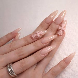 May include: Long, almond-shaped artificial nails in a nude color. Two nails feature light pink cross-shaped embellishments. A silver ring is visible on one finger. The nails are glossy and well-manicured.
