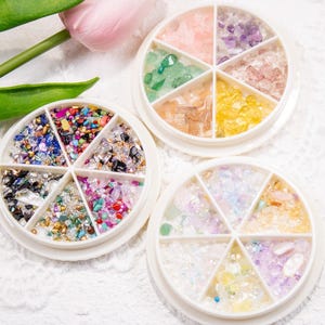 May include: Three white, circular containers filled with colorful, crushed gemstones and glitter. Each container is divided into six sections, displaying a variety of colors including pink, green, yellow, and purple. A pink tulip and green leaves are in the background.