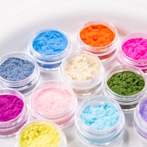 Può includere: Una collezione di piccoli vasetti di plastica trasparente riempiti di polveri vibranti e colorate. Le polveri sono in tonalità di blu, arancione, rosa, rosso, verde, giallo e viola. I vasetti sono disposti su un piatto bianco e rotondo.