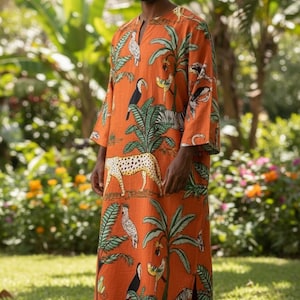 May include: An orange, long-sleeved caftan featuring a tropical print with toucans, zebras, leopards, and palm trees. The caftan has a V-neck and side slits, and is worn by a person standing in a garden.