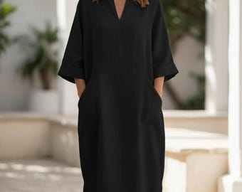 May include: A black, long-length dress with a V-neckline and elbow-length sleeves. The dress features two large front pockets and a relaxed fit. The model is standing outdoors in front of a white building.