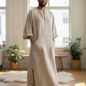 Può includere: Un caftano in lino beige con scollatura a V e tasche laterali. L'indumento a maniche lunghe arriva alle caviglie e ha spacchi laterali. La persona è in piedi a piedi nudi in una stanza ben illuminata con piante e una finestra.