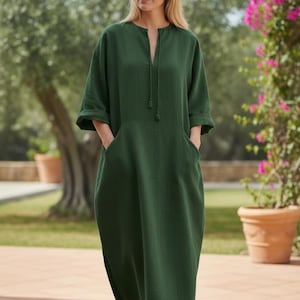 May include: A dark green, long-sleeved dress with a split neckline and two front pockets. The dress has a relaxed fit and falls to the ankles. The model is standing outside on a brick patio.