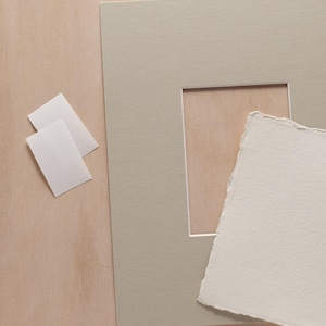 May include: A rectangular light gray picture mat with a central cutout, a piece of off-white textured paper with rough deckled edges, and two small, plain white rectangular cards arranged on a flat surface.