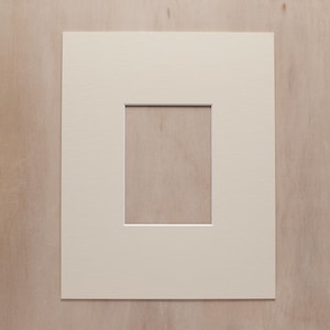 May include: A rectangular off-white paper mat board featuring a centered vertical rectangular cutout. The mat board has a smooth, matte finish and clean, straight edges, designed to frame artwork or photographs with a consistent border.