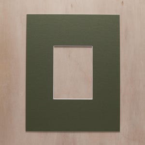 May include: A rectangular olive green picture mat board featuring a centered vertical rectangular cutout. The inner edge of the cutout is finished with a clean white border, providing a sharp contrast against the solid matte olive green surface.