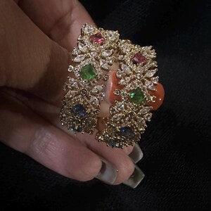 May include: A gold cuff bracelet with detailed embellishments. It features clear and coloured gemstones, including pink, green, and blue stones. The design incorporates a floral or leaf-like pattern.