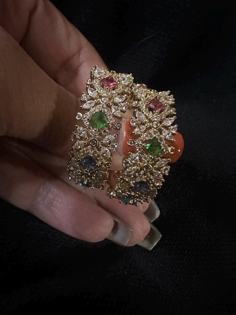 May include: A gold cuff bracelet with detailed embellishments. It features clear and coloured gemstones, including pink, green, and blue stones. The design incorporates a floral or leaf-like pattern.