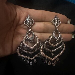May include: A pair of ornate, silver-toned chandelier earrings. Each earring features a diamond-shaped top with a leaf design, followed by layered, heart-shaped tiers, and teardrop-shaped dangles. The earrings are set with clear, sparkling stones.